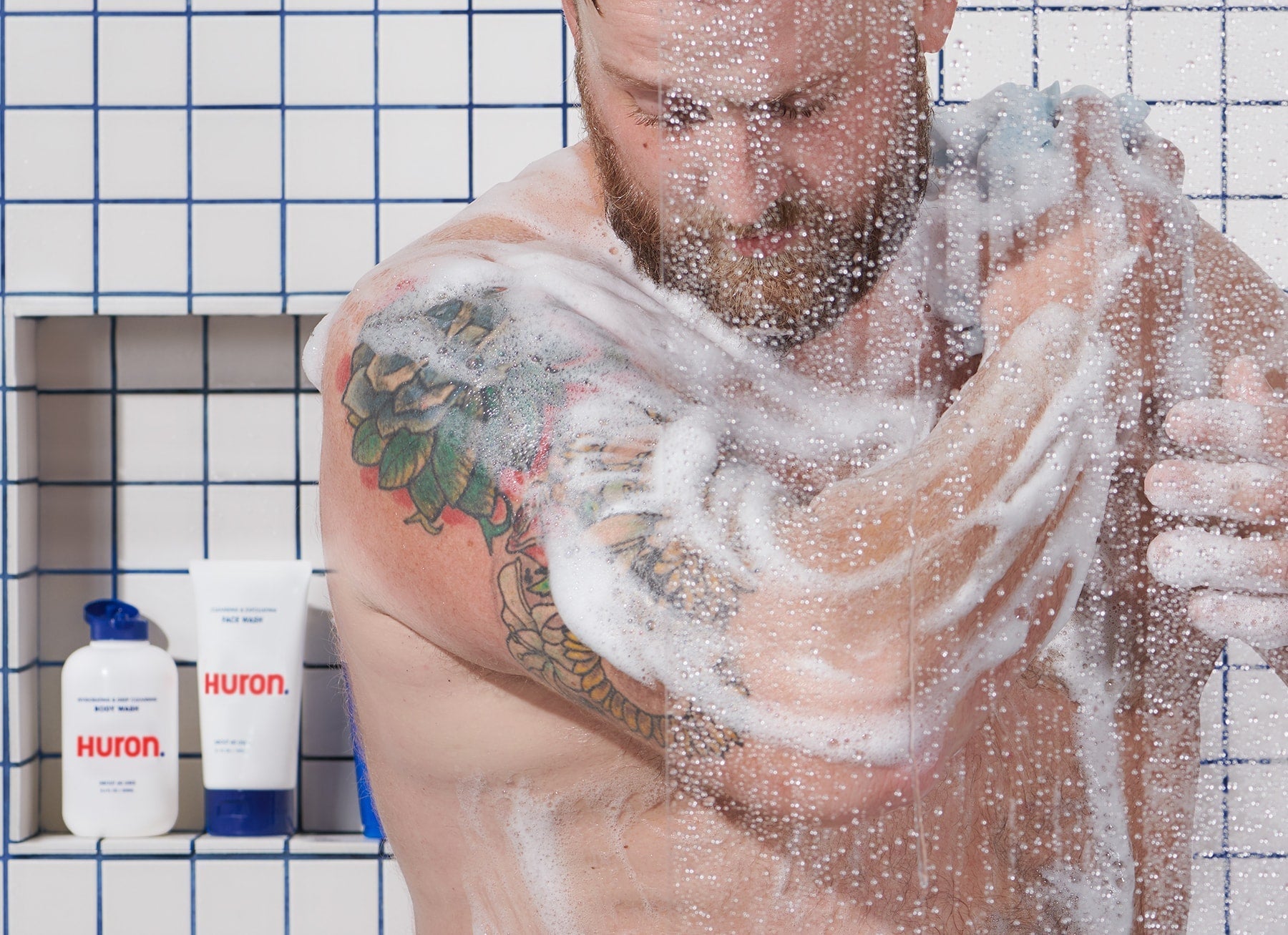 Bearded man lathering in tiled shower, tattooed shoulder visible; HURON. FACE WASH bottle on shelf.