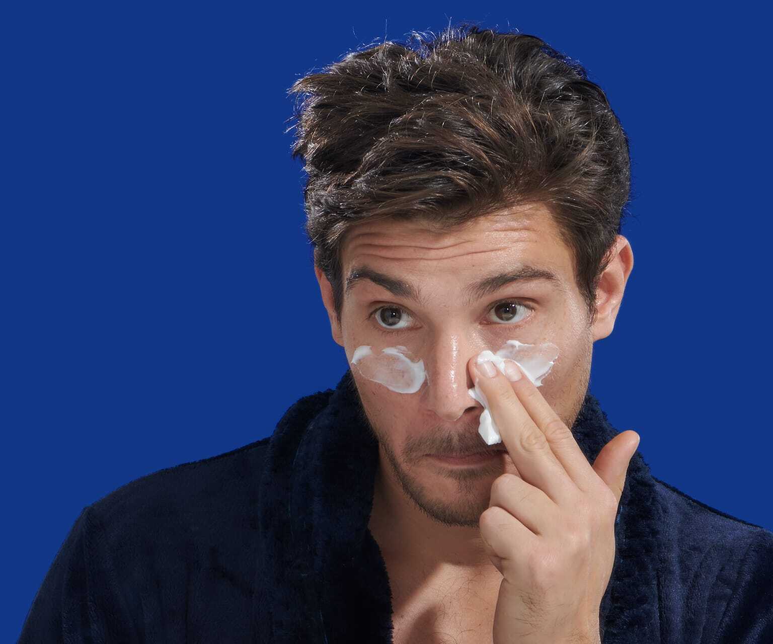 Young man in dark robe applying white eye cream under both eyes against a solid blue background.