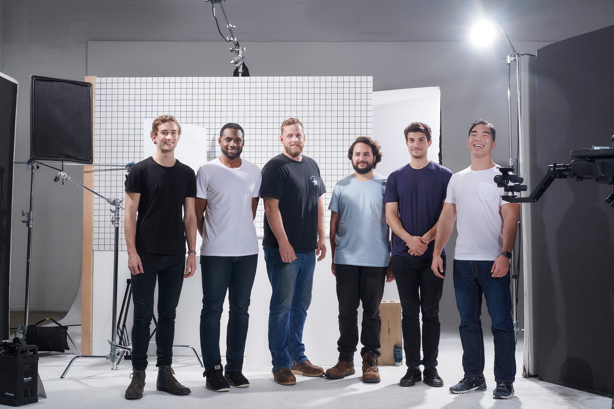 Six men standing in a photography studio with lighting equipment and a grid backdrop, smiling and facing the camera.
