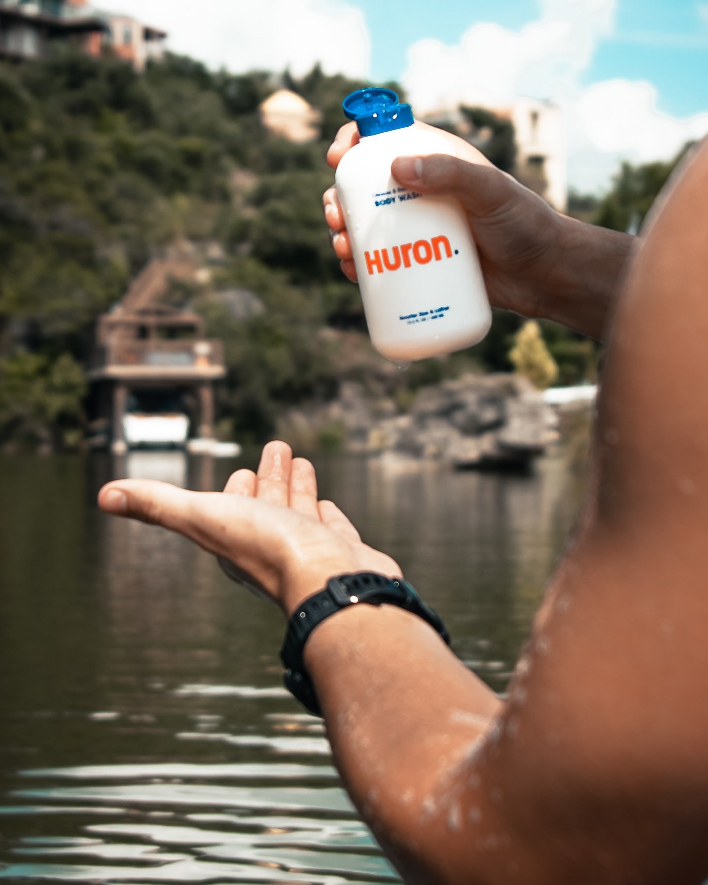 Person holding bottle labeled Huron. and BODY WASH, pouring into open hand over a lake