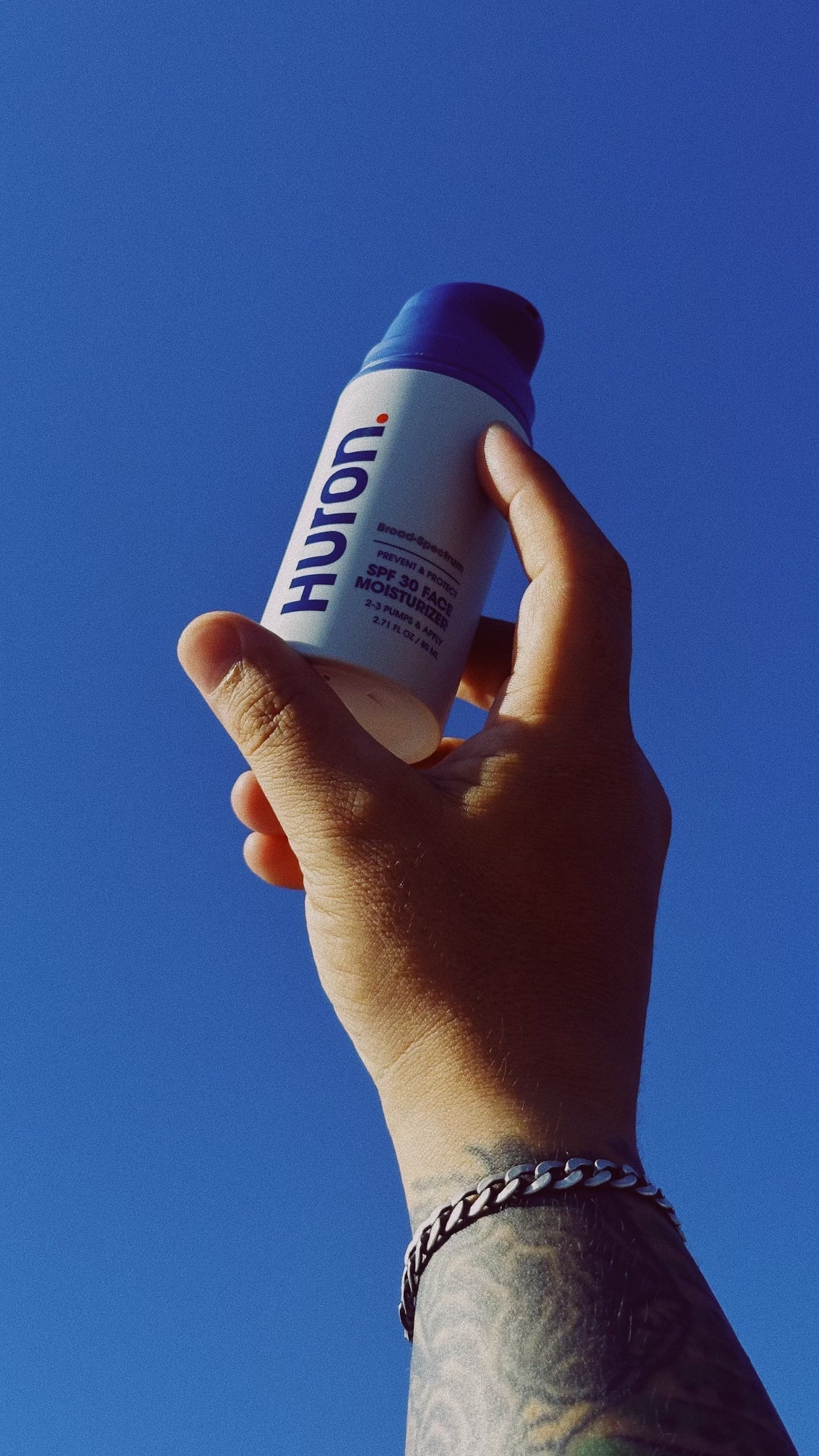 Hand holding a bottle of Huron Broad-Spectrum SPF 30 Face Moisturizer against a clear blue sky. Visible text: "Huron. Broad-Spectrum PREVENT & PROTECT SPF 30 FACE MOISTURIZER 2-3 PUMPS & APPLY 2.7 FL OZ / 80 ML".