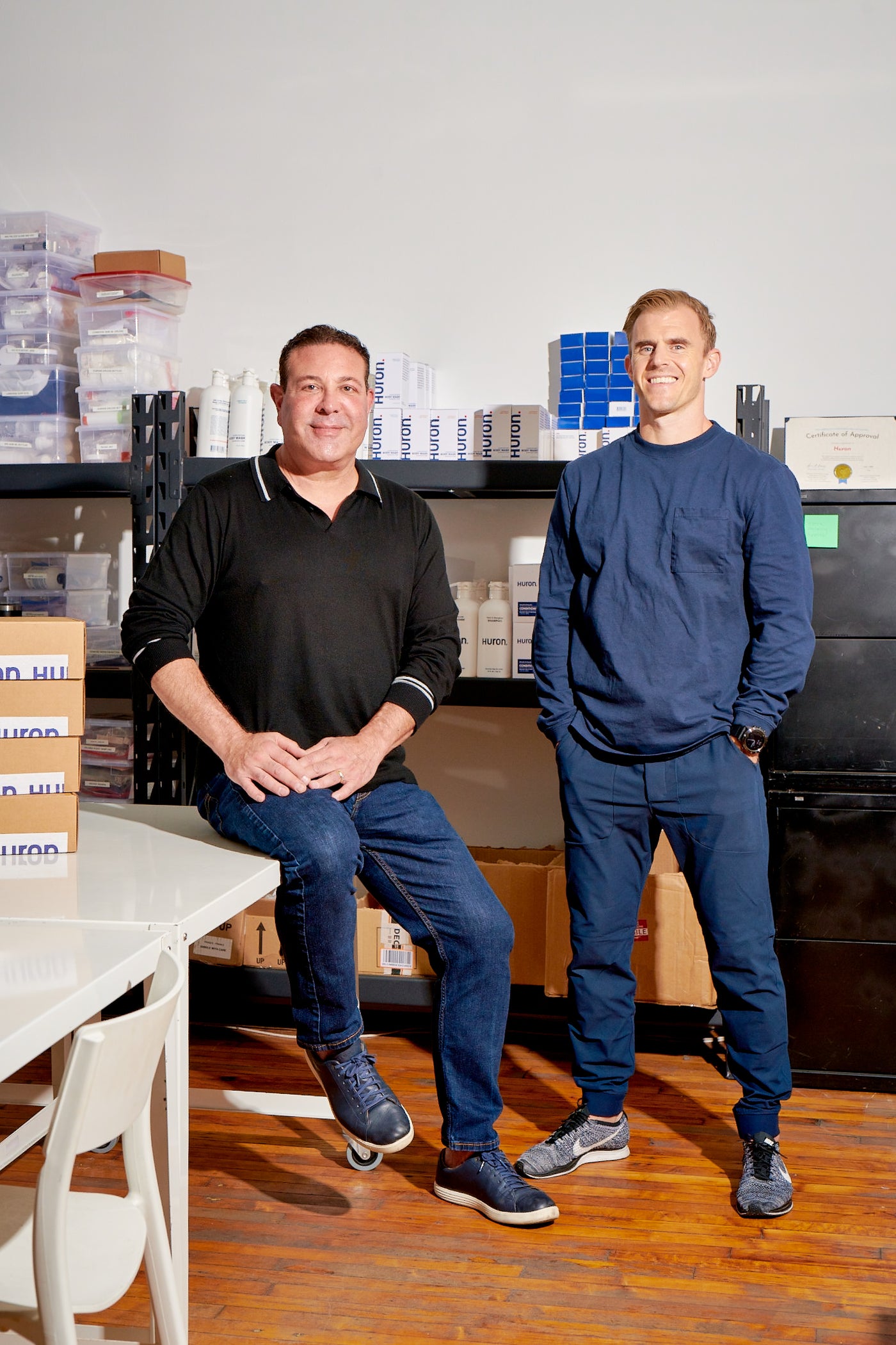 Two men standing in a workspace with personal care products in the background.