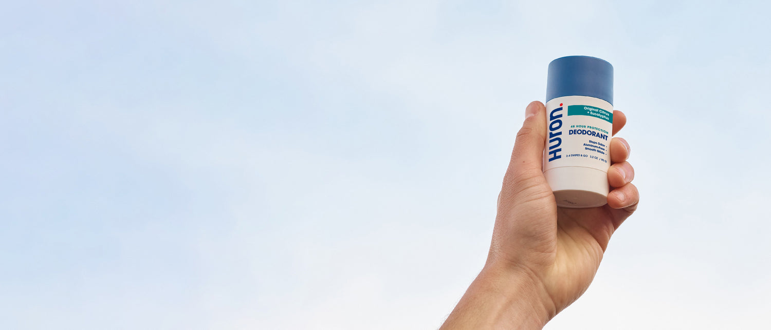 Hand holding Huron deodorant with blue cap against a clear sky background. Text on bottle: 'HURON DEODORANT'.