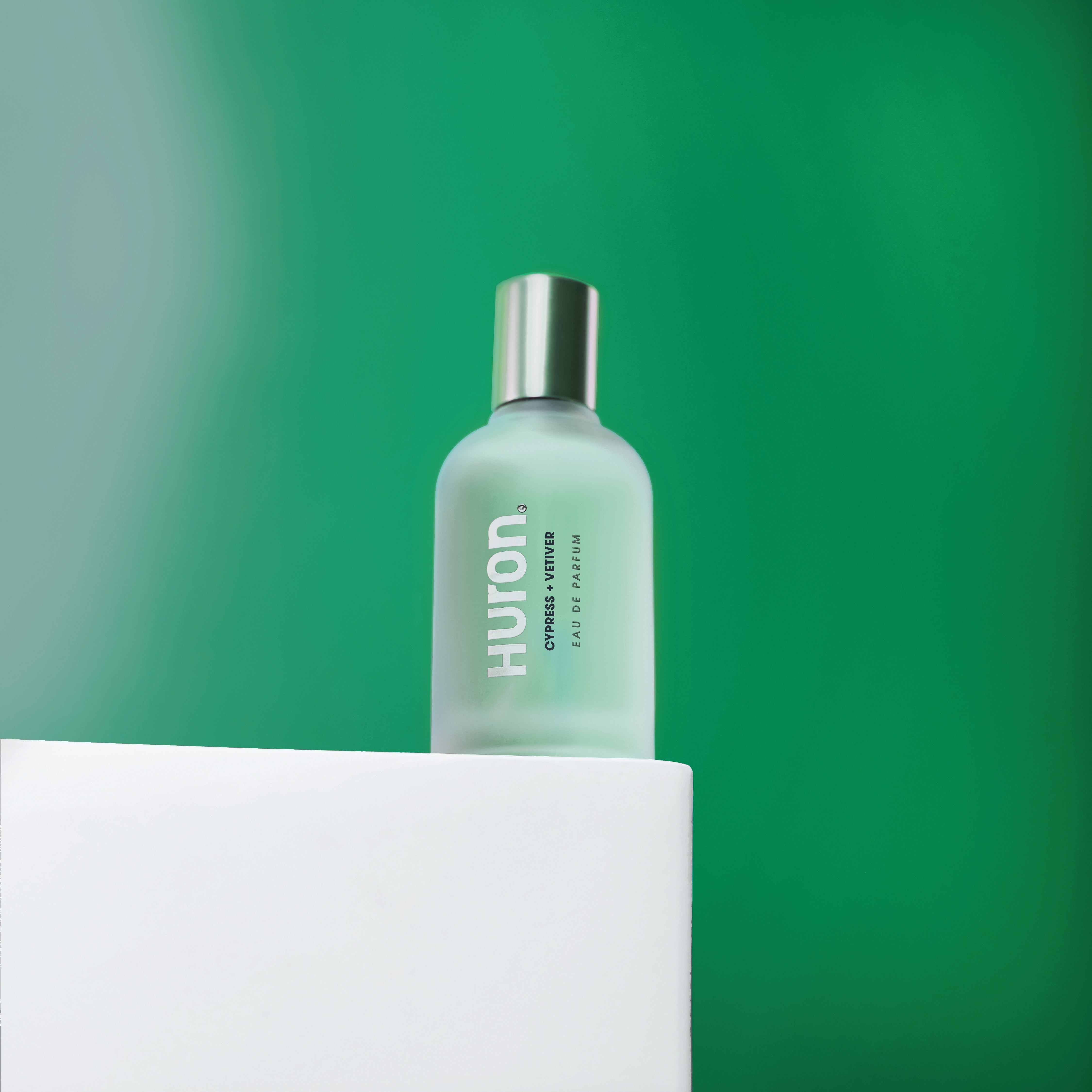 Huron Cypress + Vetiver Eau de Parfum bottle with frosted glass and silver cap on white surface against green background #scent_cypress + vetiver