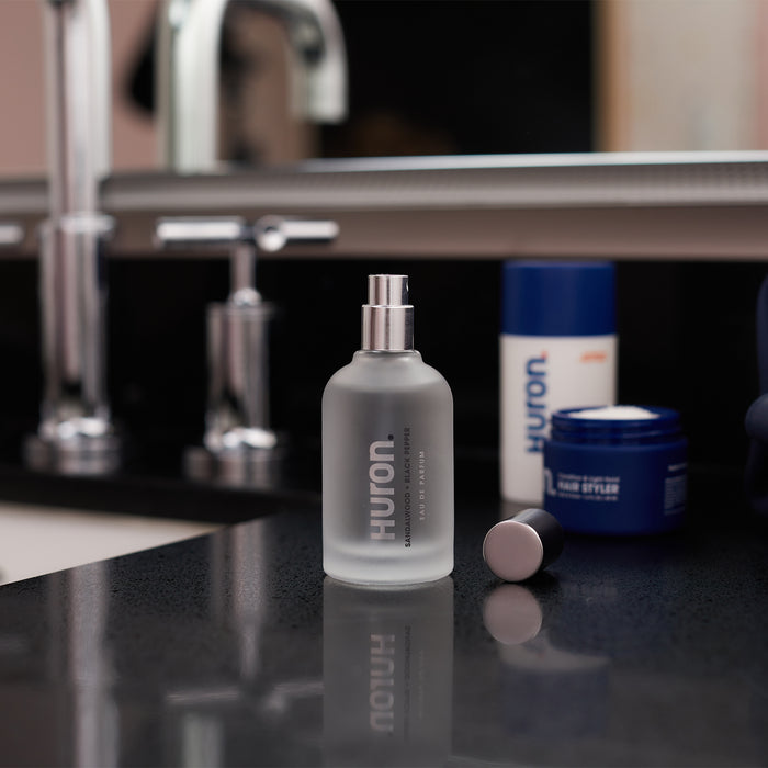 Frosted glass Huron spray bottle with its cap beside it on a bathroom counter, blue Huron containers and mirror in background