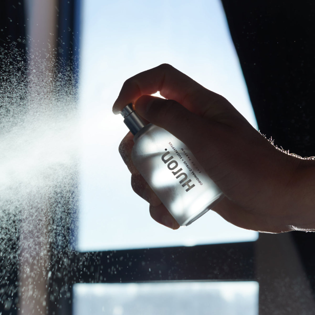 A person spraying Huron cologne from a bottle, with mist visible in the air.