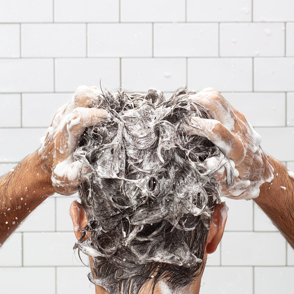 Back of a person’s head with wet hair being lathered with shampoo in a tiled shower