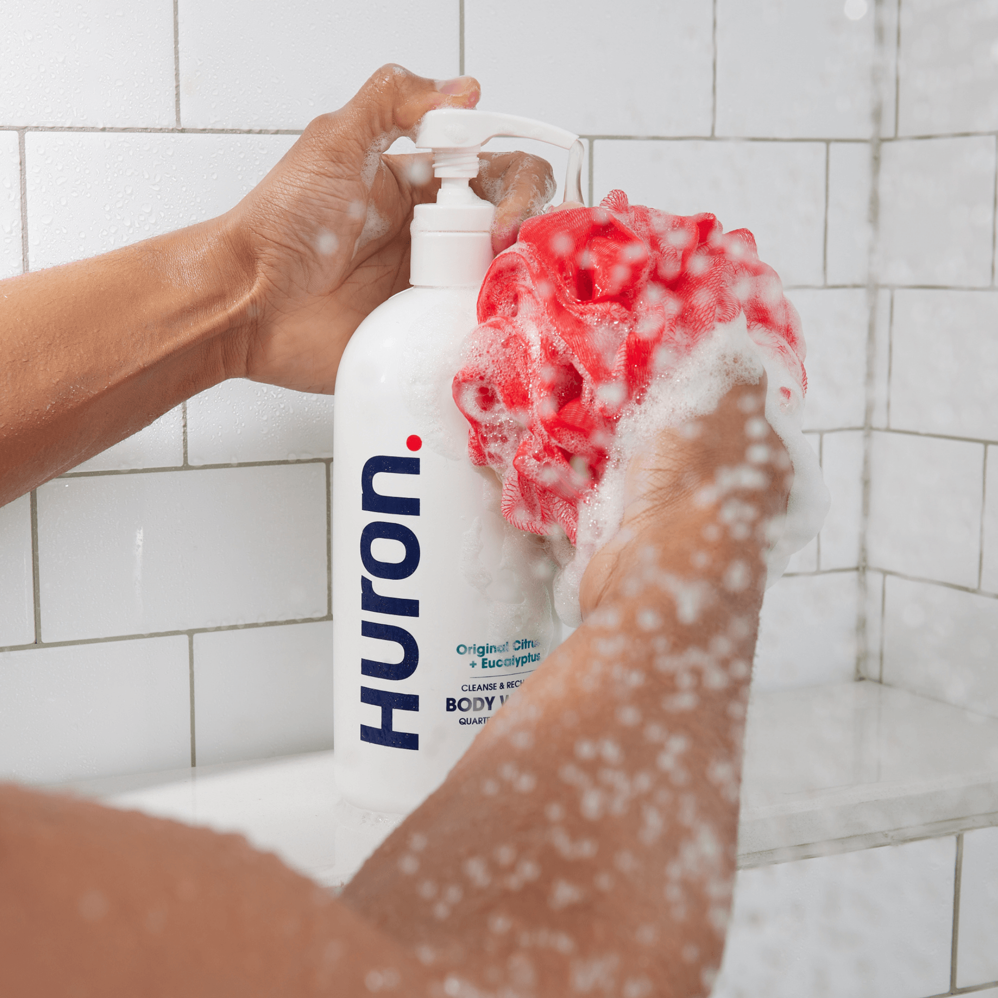 Hands pressing a Huron pump bottle to dispense body wash onto a red bath sponge in a tiled shower