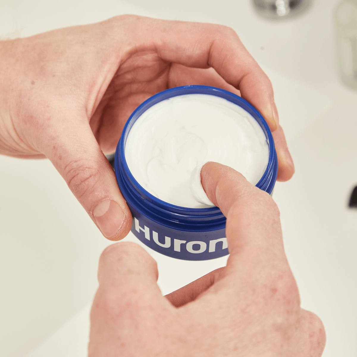 Hands scooping white styling cream from an open blue jar labeled Huron.