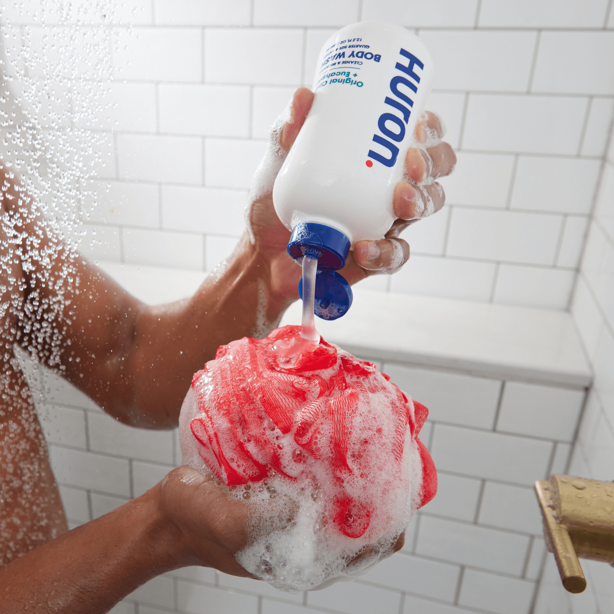 Hands pouring Huron body wash onto a red shower loofah in a white-tiled shower