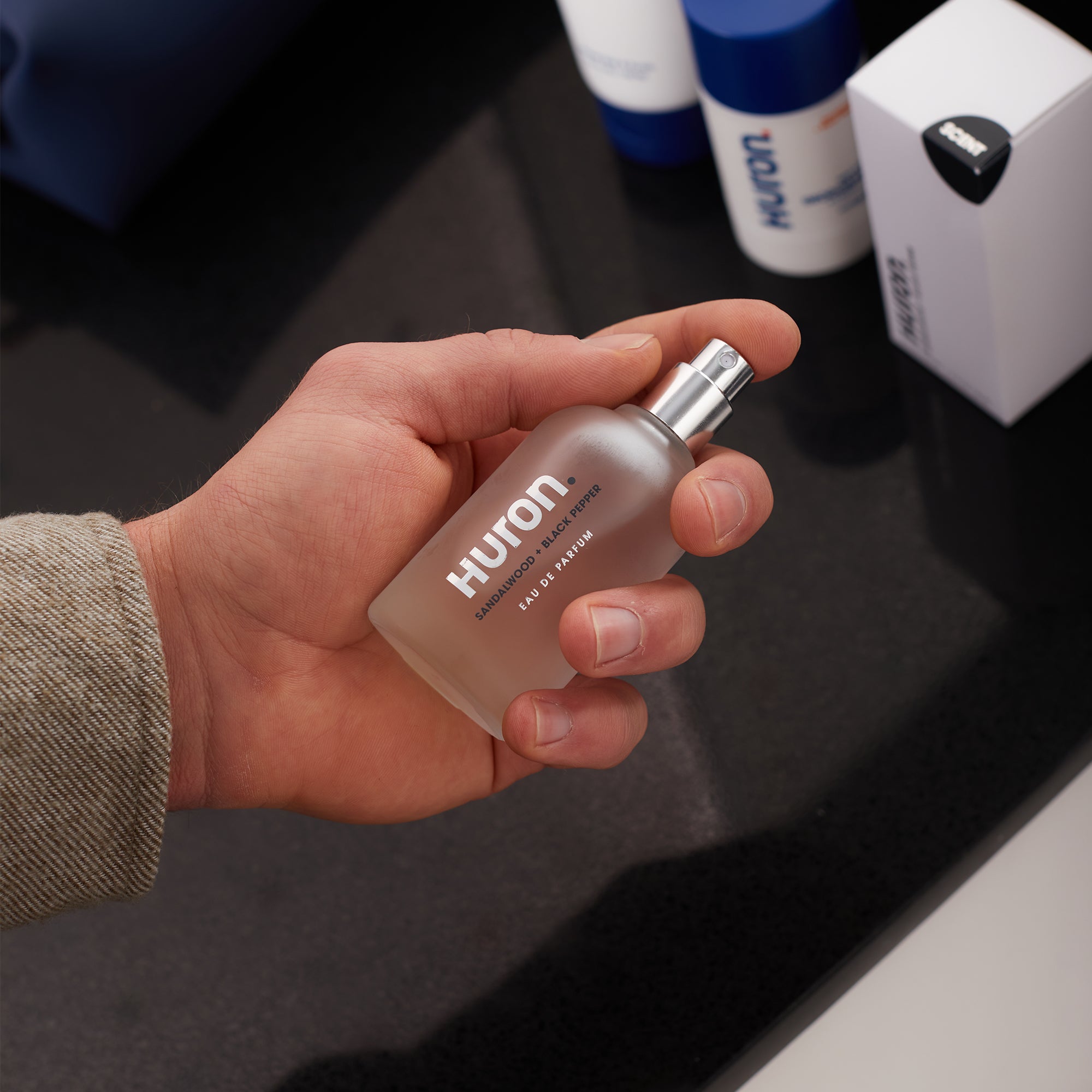 Hand holding a frosted glass spray bottle labeled 'Huron Eau de Parfum' with a silver spray nozzle.