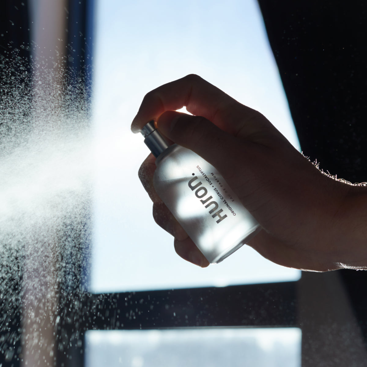 Hand-held view of Huron Eau de Parfum bottle showcasing spray mist and frosted glass with a bright backlit window background