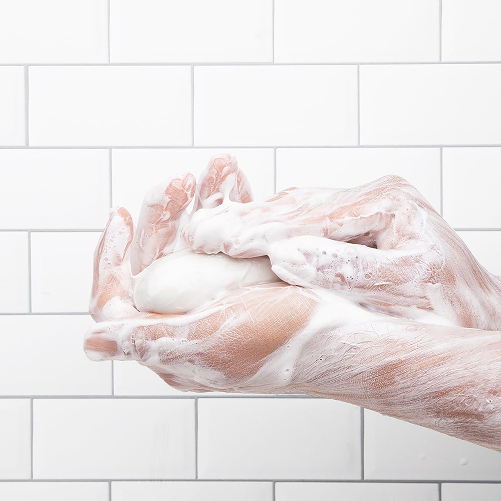 Hands covered in soap lather holding a white bar of soap against a white tiled background.