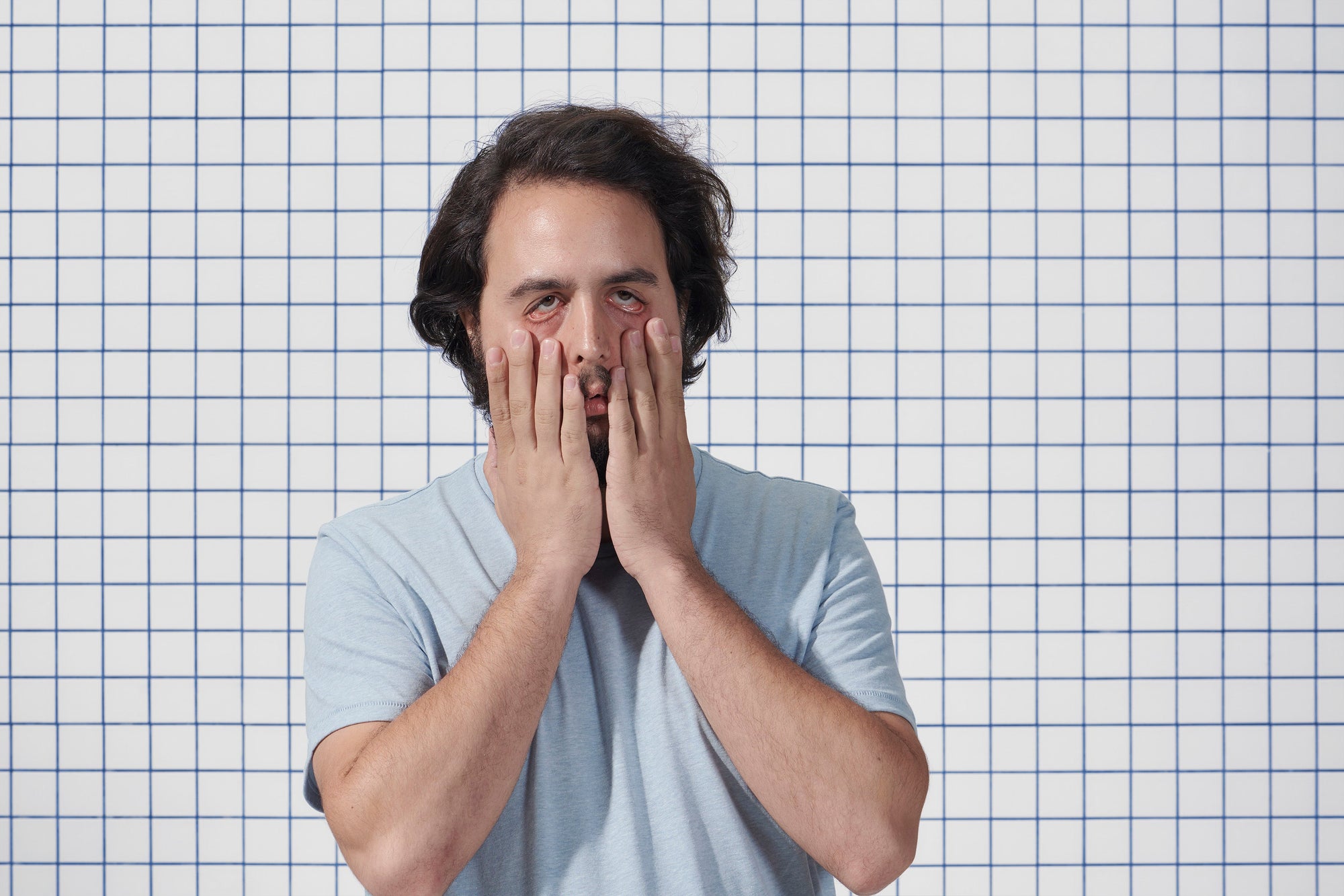 Man pressing his face with hands, appearing tired or stressed, standing against a white wall with a blue grid pattern.