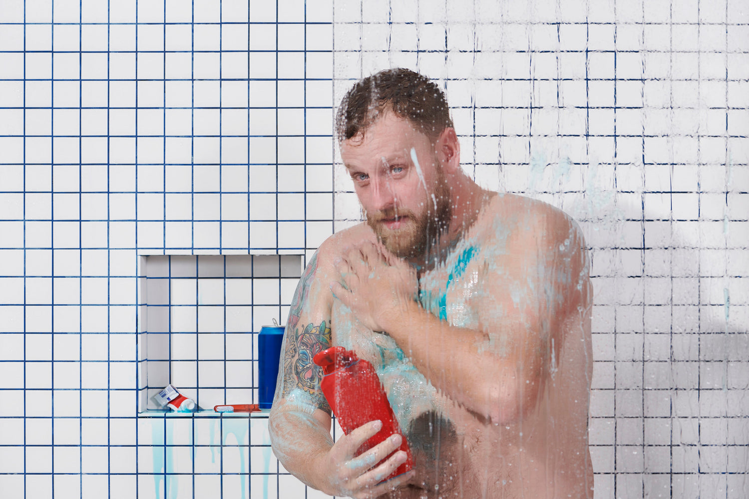 Man with tattoos in a shower holding a red bottle, with blue paint or soap on his body and wet glass in front.