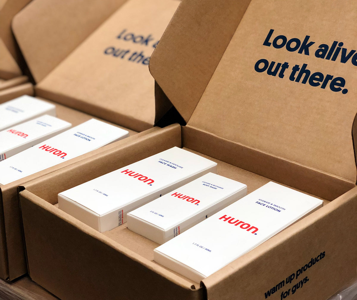 Open cardboard box containing Huron skincare products labeled Cleanse & Exfoliate Face Wash, Cleanse & Exfoliate Body Wash, and Hydrate & Smooth Face Lotion. Text on box flap reads 'Look alive out there.'