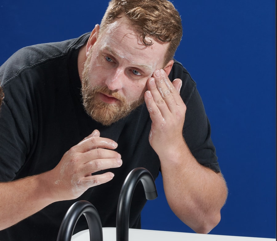 Bearded man applying facial cream near a sink with black faucets against a blue background.
