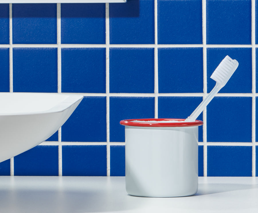 White toothbrush in a white cup with a red rim on a white surface, blue tiled wall background.