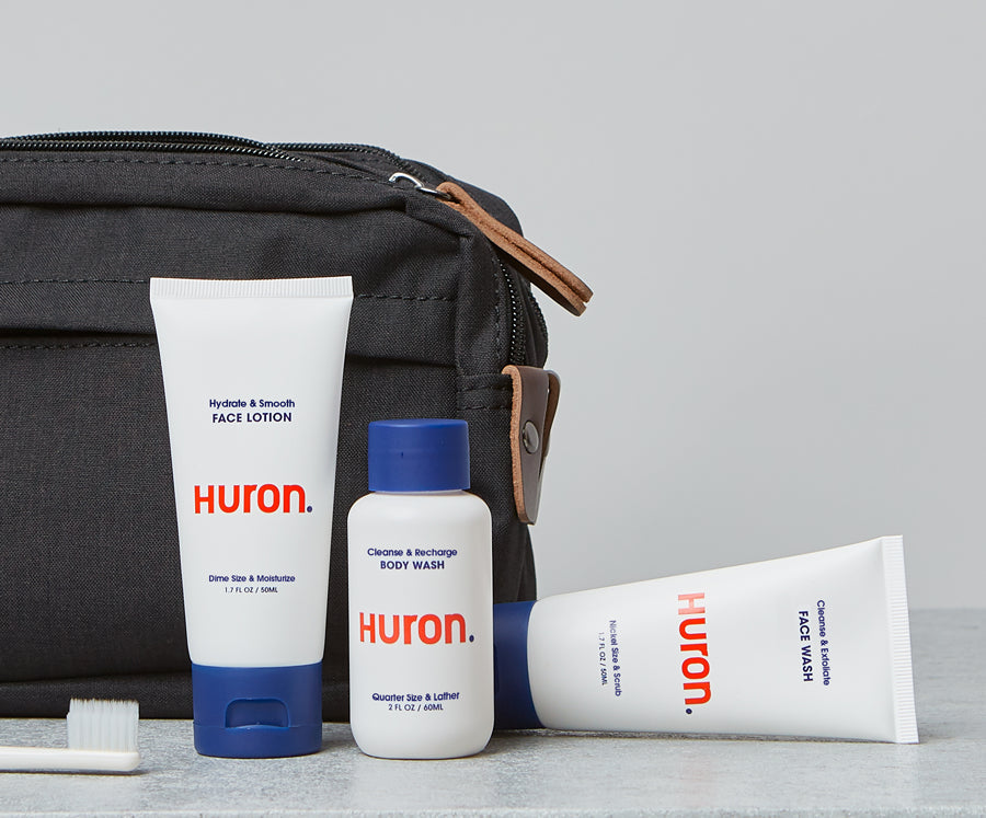 Travel toiletry bag with Huron brand face lotion, body wash, face wash, and a toothbrush on a gray surface.
