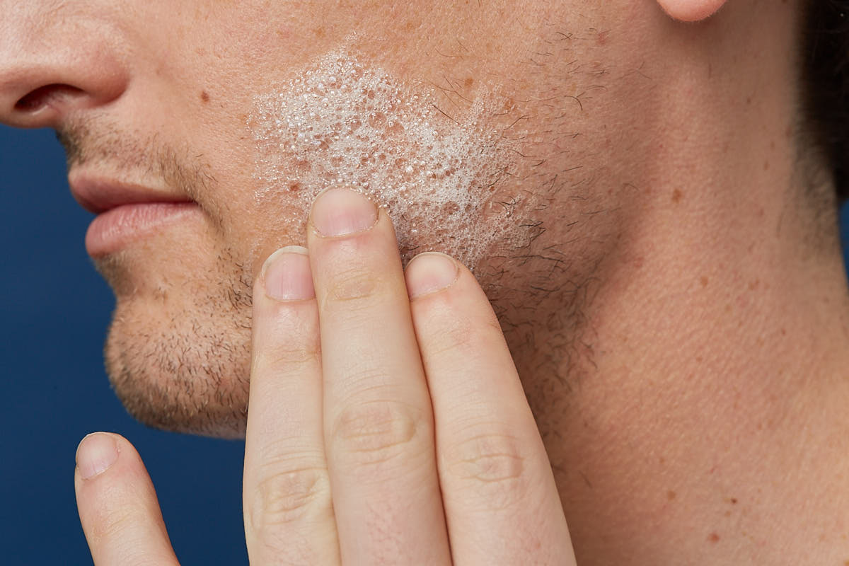 Close-up of a man applying foamy facial cleanser to his cheek with fingers against a blue background