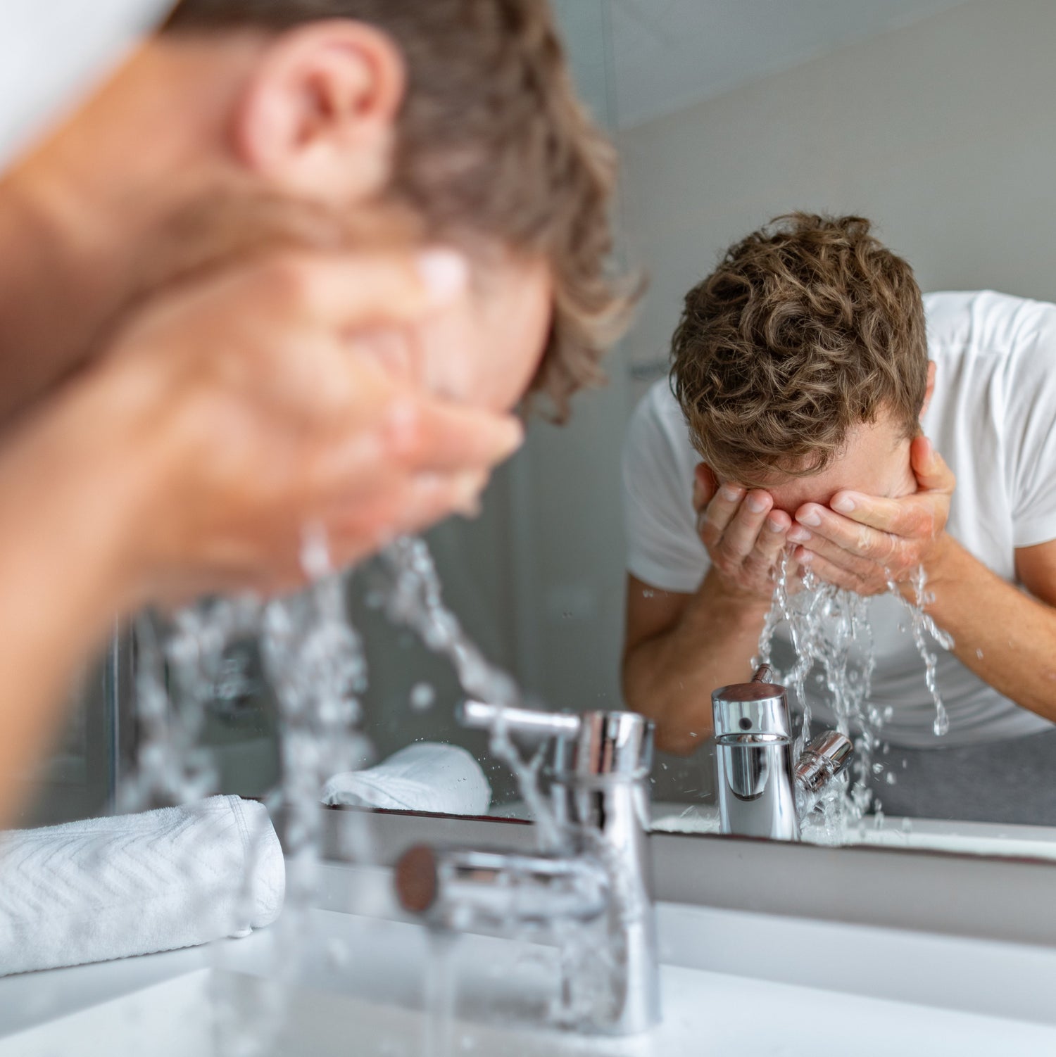 Man washing his face with water at a bathroom sink, reflected in the mirror.