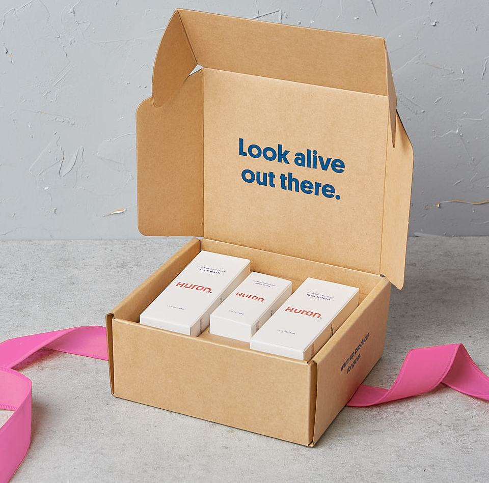 Cardboard box with three Huron skincare product boxes inside. Inside lid text reads 'Look alive out there.' Pink ribbon beside the box.