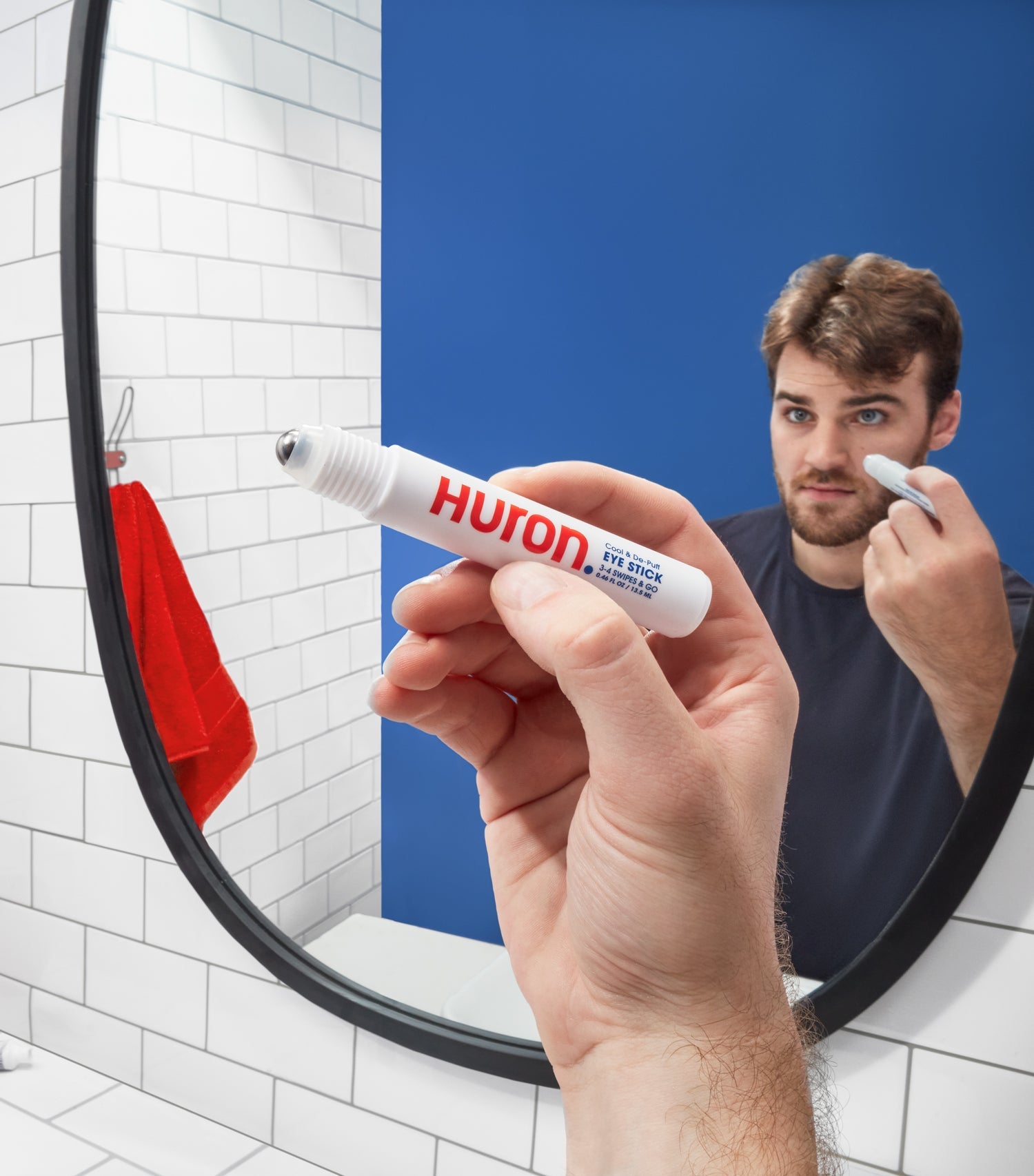 Man applying Huron Cool & De-Puff Eye Stick with metal rollerball applicator in front of a bathroom mirror, visible text: 'Huron', 'Cool & De-Puff', 'EYE STICK', '3-4 SWIPES & GO', '0.46 FL OZ / 13.5 ML'
