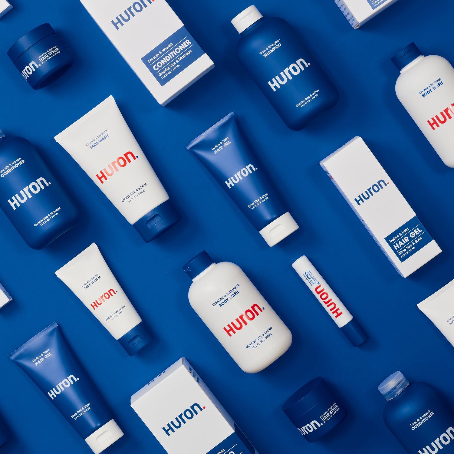 Flat lay of Huron skincare and grooming products showcasing blue-and-white bottles, tubes, and boxes on a solid blue background