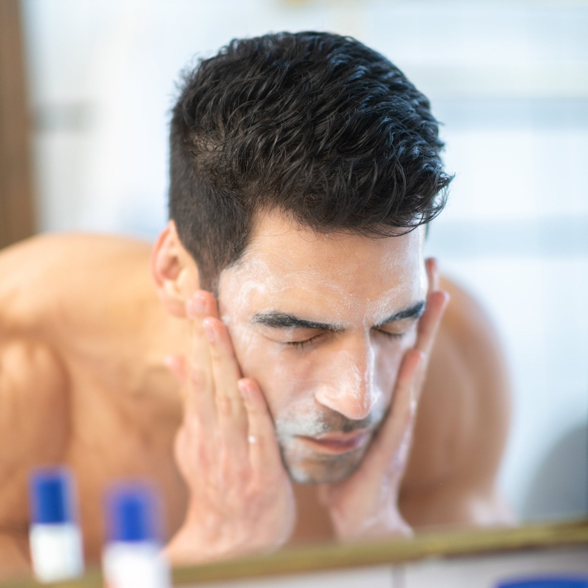Man washing his face with foam in front of a bathroom mirror as part of a morning routine