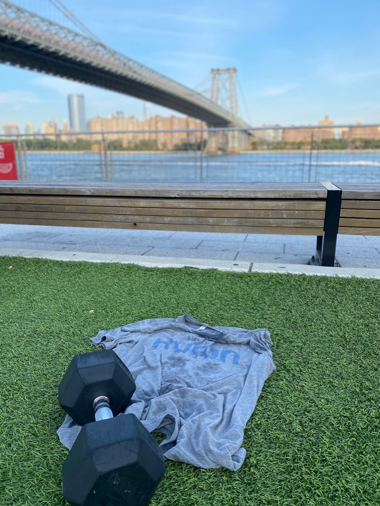 Gray workout shirt with partially visible blue text and a black dumbbell on green grass near a wooden bench and a bridge over water in the background.