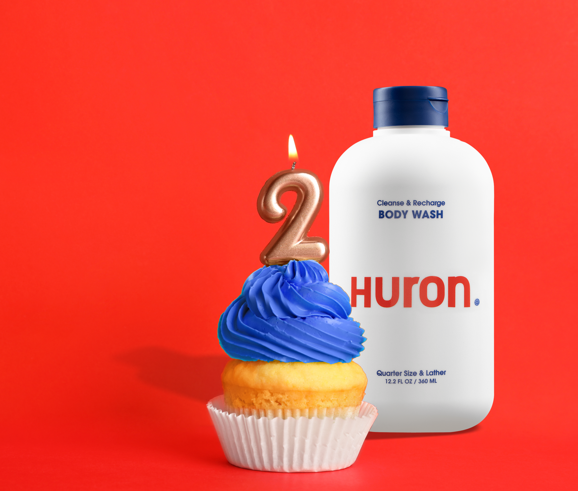 Cupcake with blue frosting and lit number 2 candle next to a white bottle labeled 'Cleanse & Recharge BODY WASH Huron. Quarter Size & Lather 12.2 FL OZ / 360 ML'