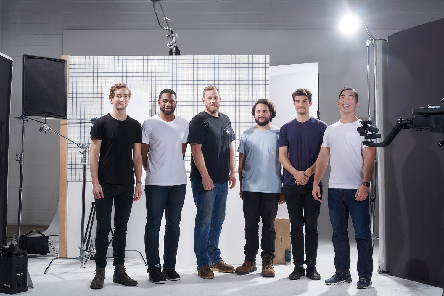 Six men standing in a photography studio with lighting equipment and a grid backdrop, smiling and facing the camera.