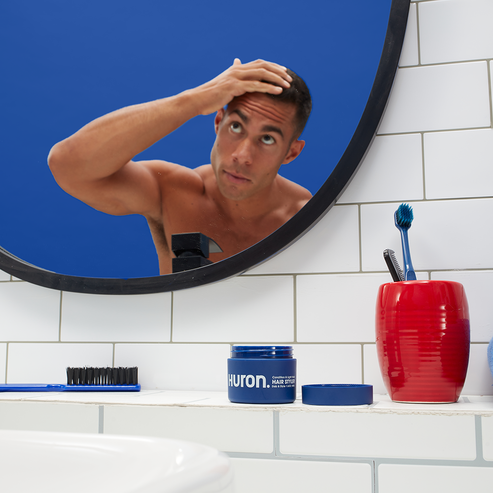 Man looking at his hair in a bathroom mirror with grooming products including a Huron hair styler, toothbrush, and comb on the counter