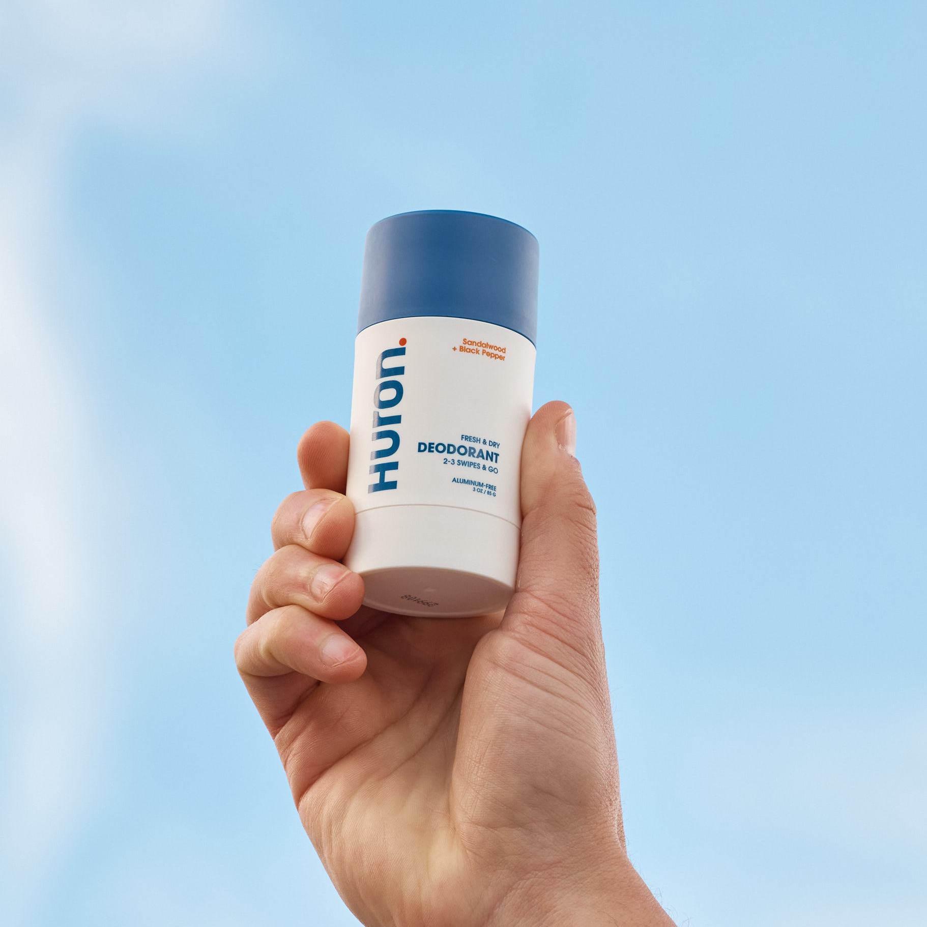 A hand holding Huron deodorant stick against a blue sky.