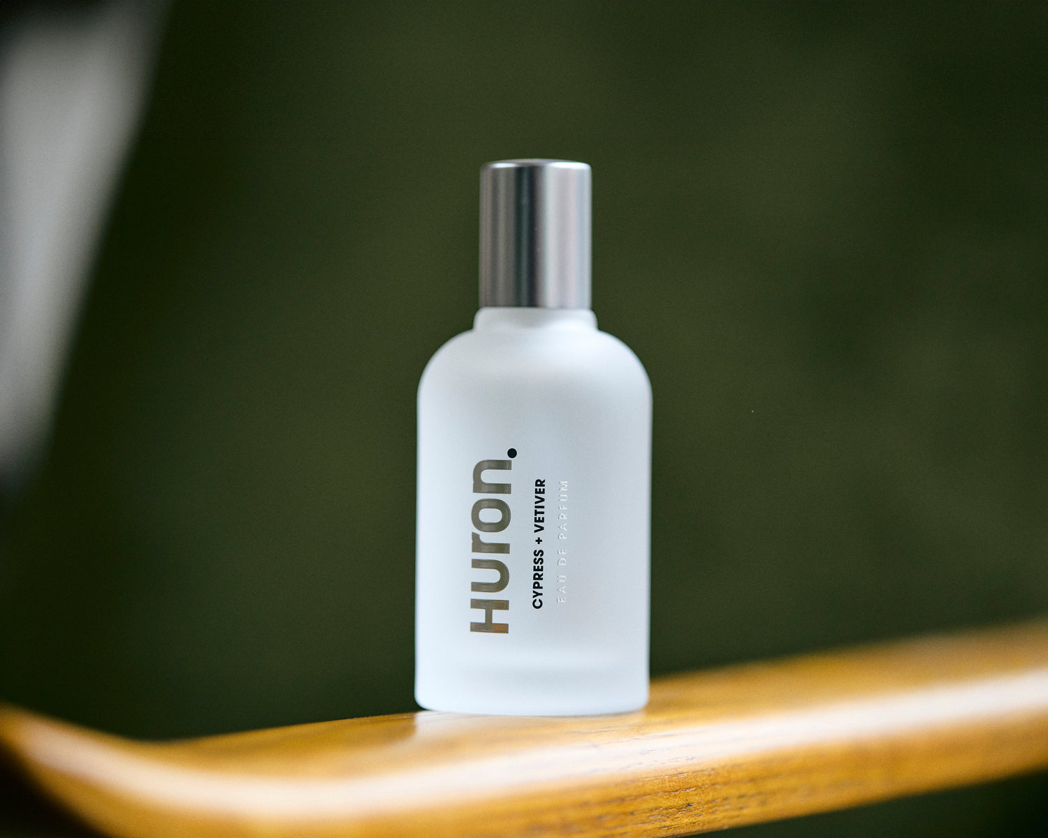 Frosted glass perfume bottle labeled 'Huron. CYPRESS + VETIVER EAU DE PARFUM' on a wooden surface with a blurred green background