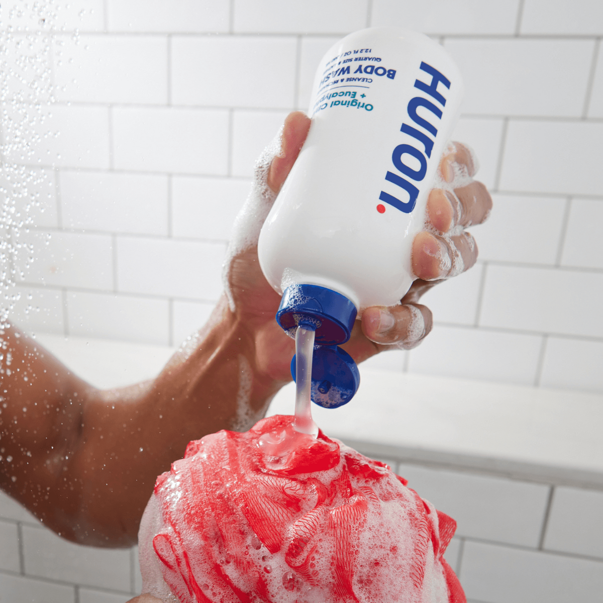 A person pouring body wash from a bottle onto a loofah in a shower.