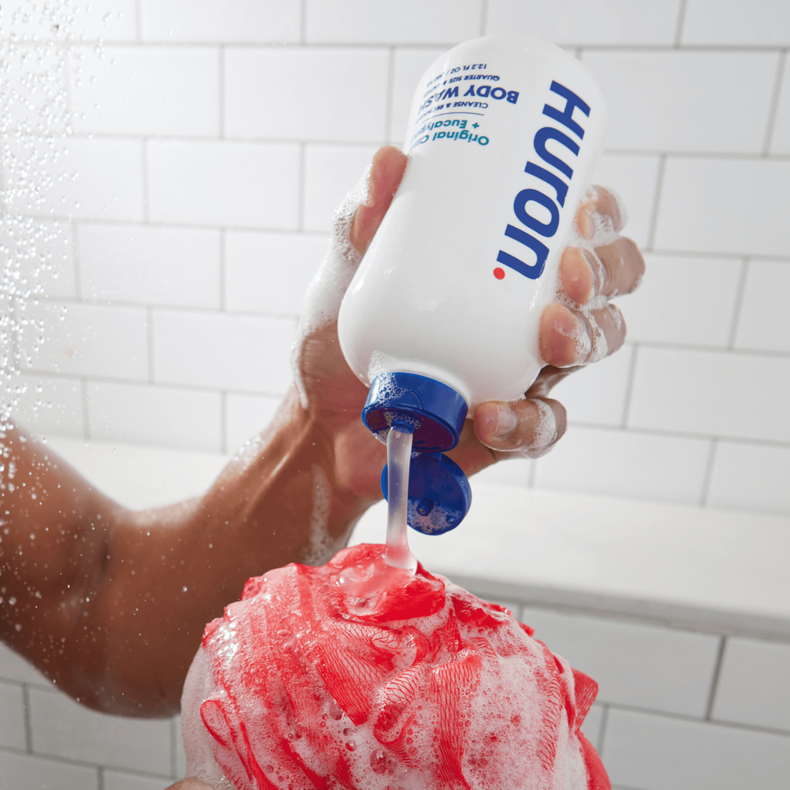 A person pouring body wash from a bottle onto a loofah in a shower.