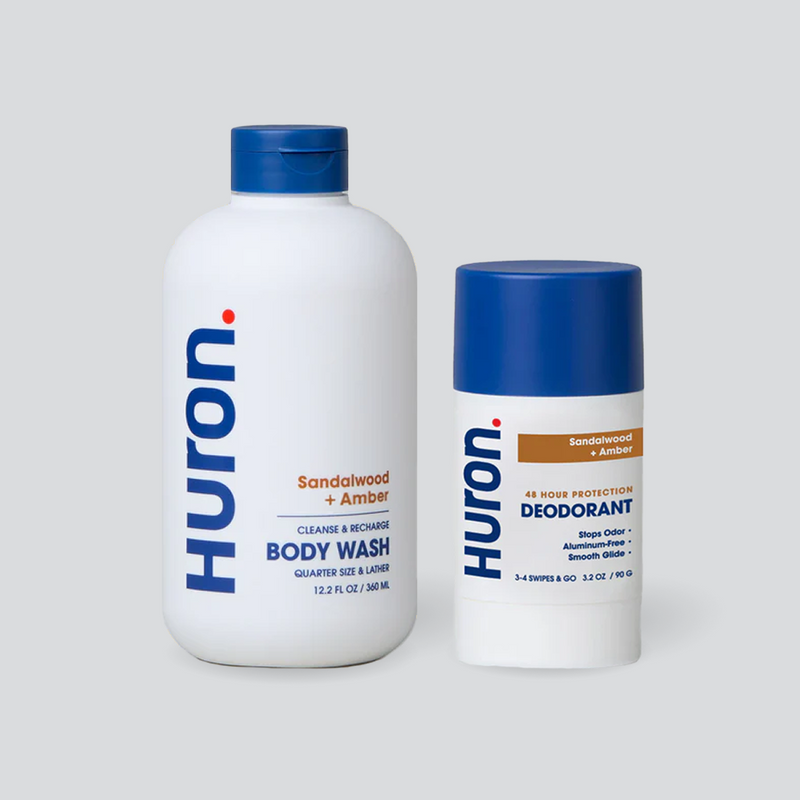 Two Huron personal care products: a body wash labeled 'Sandalwood + Black Pepper' and a deodorant labeled 'Sandalwood + Amber'. Both have blue caps and white bottles with visible text including 'Huron', 'Cleanse & Recharge', 'Body Wash', '48 Hour Protection', 'Stops Odor', 'Aluminum-Free', and 'Smooth Glide'. #scent_sandalwood + amber