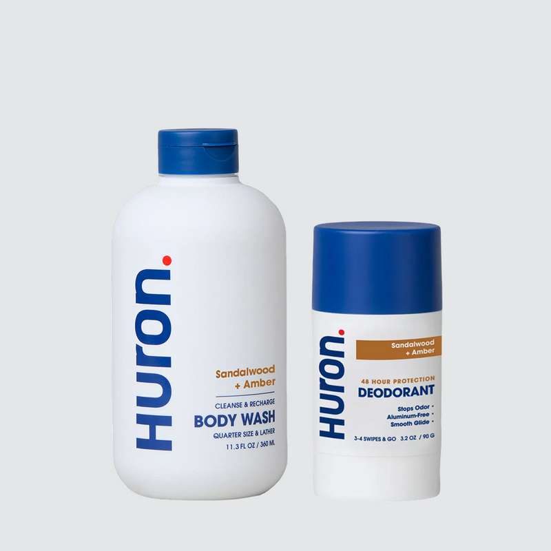 Two Huron personal care products: a body wash labeled 'Sandalwood + Black Pepper' and a deodorant labeled 'Sandalwood + Amber'. Both have blue caps and white bottles with visible text including 'Huron', 'Cleanse & Recharge', 'Body Wash', '48 Hour Protection', 'Stops Odor', 'Aluminum-Free', and 'Smooth Glide'. #scent_sandalwood + amber