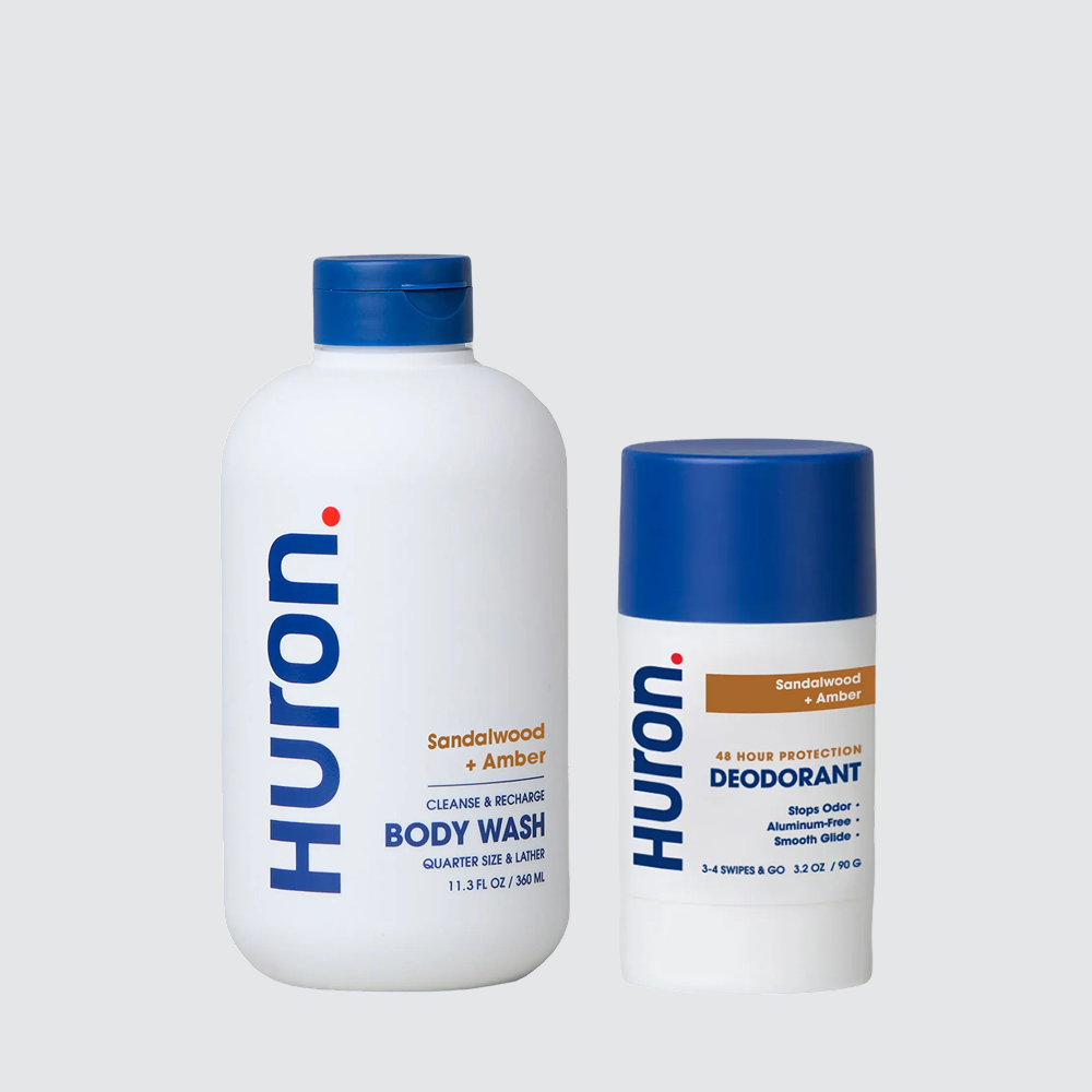 Two Huron personal care products: a body wash labeled 'Sandalwood + Black Pepper' and a deodorant labeled 'Sandalwood + Amber'. Both have blue caps and white bottles with visible text including 'Huron', 'Cleanse & Recharge', 'Body Wash', '48 Hour Protection', 'Stops Odor', 'Aluminum-Free', and 'Smooth Glide'. #scent_sandalwood + amber