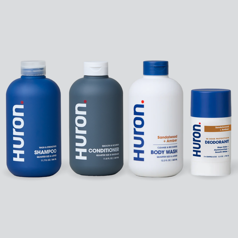 Four HURON toiletry bottles labeled HURON: SHAMPOO, CONDITIONER, BODY WASH (Sandalwood + Black Pepper), and DEODORANT. #scent_sandalwood + amber