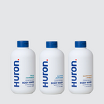 Front view of Huron body wash trio: three white bottles with blue caps labeled Citrus + Eucalyptus; Sea Salt + Driftwood; Sandalwood + Amber