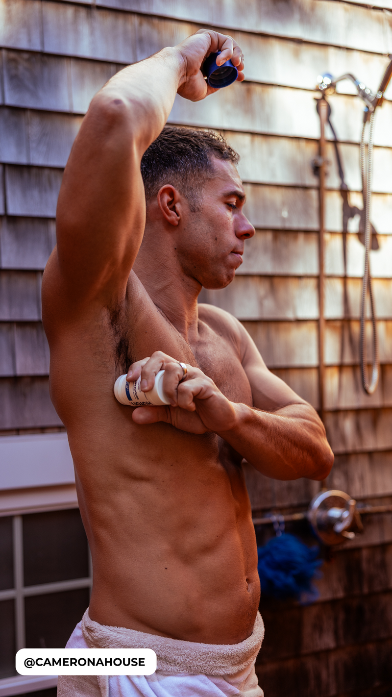 Male in towel applying stick deodorant to his right armpit, visible social tag '@CAMERONAHOUSE'.