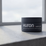 Front view of Huron Hair Clay jar showcasing product label and matte packaging on a concrete windowsill lit by soft window light