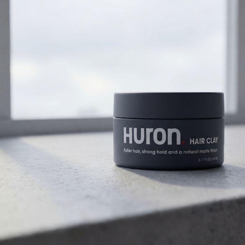 Front view of Huron Hair Clay jar showcasing product label and matte packaging on a concrete windowsill lit by soft window light