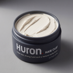 Angled top view of Huron Hair Clay jar open showing creamy matte texture and label, on neutral gray background