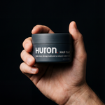 Close-up of a hand holding Huron Hair Clay jar showing the label, matte black packaging and 2.77 fl oz against a dark background