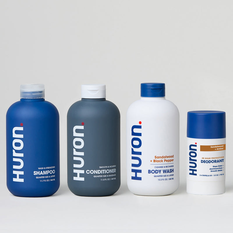 Four HURON toiletry bottles labeled HURON: SHAMPOO, CONDITIONER, BODY WASH (Sandalwood + Black Pepper), and DEODORANT. #scent_sandalwood + amber