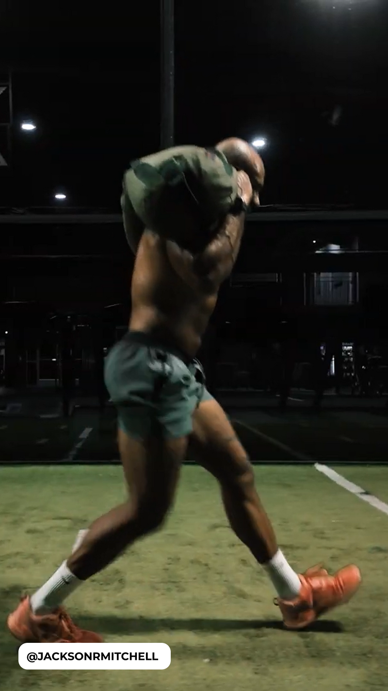 Athlete carrying a sandbag on his shoulder during a nighttime turf workout, in shorts and orange shoes, @JACKSONRMITCHELL