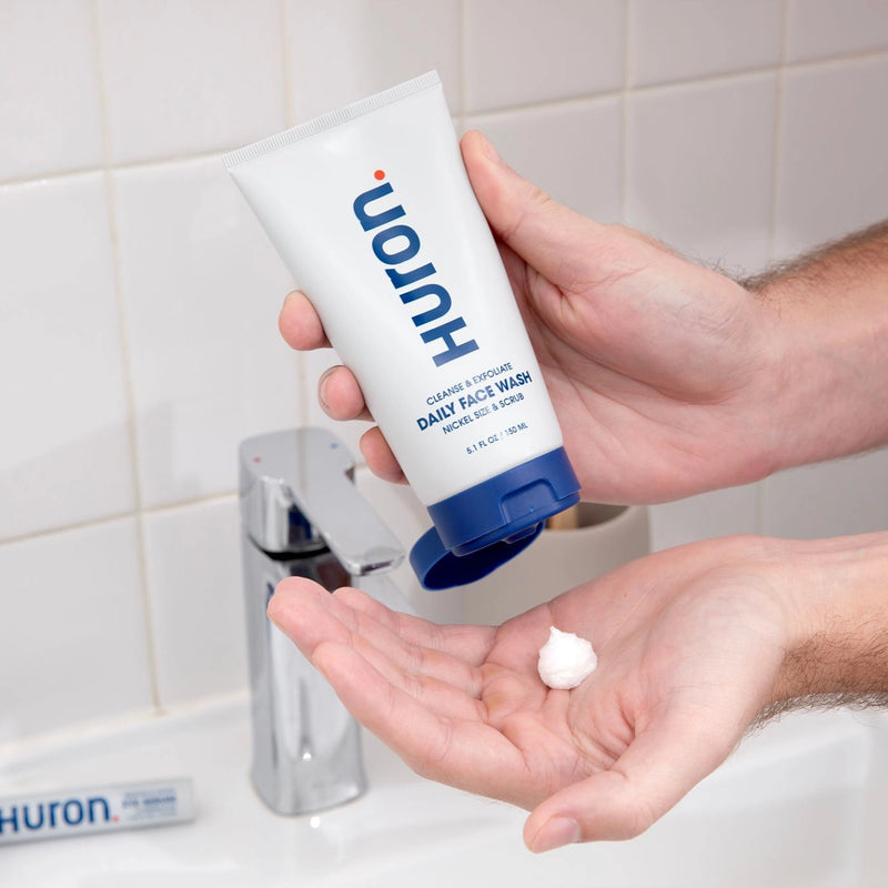 Hands hold Huron tube dispensing dollop; text: Huron CLEANSE & EXFOLIATE DAILY FACE WASH NICKEL SIZE & SCRUB 5.1 FL OZ 150 ML